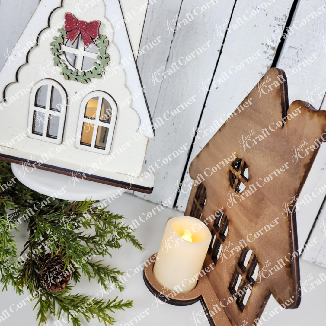 Janet's Craft Corner Victorian Gingerbread House Votive Holder DIY kit sits on a white wooden background with a flameless candle, festive greenery, and Gingerbread House Mini Shelf Sitters for seasonal home decor.