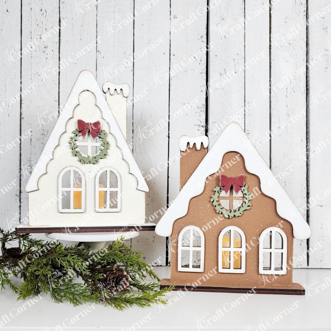 Create charming festive decor with two Victorian Gingerbread House Votive Holders from Janet's Craft Corner DIY kit—one white and one brown, each featuring snow-like rooftops, wreaths, arched windows, and pine branches.