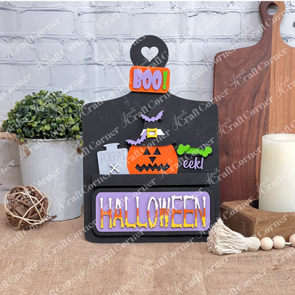 This delightful DIY home decor kit features Janet's Craft Corner's Halloween Pumpkin Interchangeable Insert, showcased on a black breadboard with a cheerful pumpkin wearing a witch hat. The vibrant "HALLOWEEN" and "BOO!" text enhance its festive appeal. Accompanied by a plant, candle, and cutting boards set against a brick wall backdrop, it's ideal for those who enjoy interchangeable craft inserts.