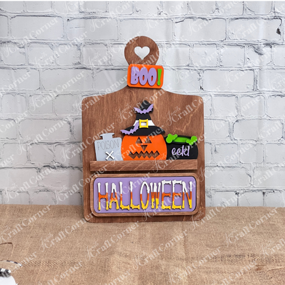 This delightful DIY home decor kit features Janet's Craft Corner's Halloween Pumpkin Interchangeable Insert, showcased on a stained breadboard  with a cheerful pumpkin wearing a witch hat. The vibrant "HALLOWEEN" and "BOO!" text enhance its festive appeal. Accompanied by a plant, candle, and cutting boards set against a brick wall backdrop, it's ideal for those who enjoy interchangeable craft inserts.