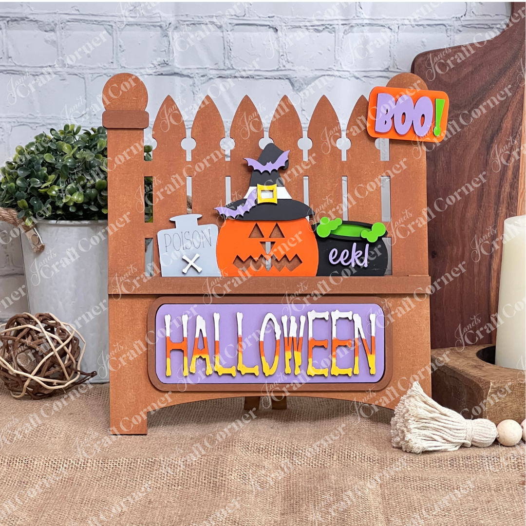 This delightful Halloween decor from Janet's Craft Corner features a wooden fence backdrop with interchangeable inserts: a pumpkin, witch's hat, "Poison" bottle, and bubbling cauldron. A cheerful "BOO!" sign hangs above, while vibrant letters spell "HALLOWEEN" at the base.