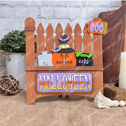 This delightful Halloween decor from Janet's Craft Corner features a wooden fence backdrop with interchangeable inserts: a pumpkin, witch's hat, "Poison" bottle, and bubbling cauldron. A cheerful "BOO!" sign hangs above, while vibrant letters spell "HALLOWEEN" at the base.