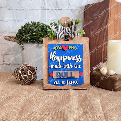 Janet’s Craft Corner Breadboard Sign Holder Shelf Sitter, a DIY farmhouse decor kit for switchable wooden signs, is shown with a “Happiness made with one roll at a time” sign, bow, greenery accents, and rustic burlap styling.