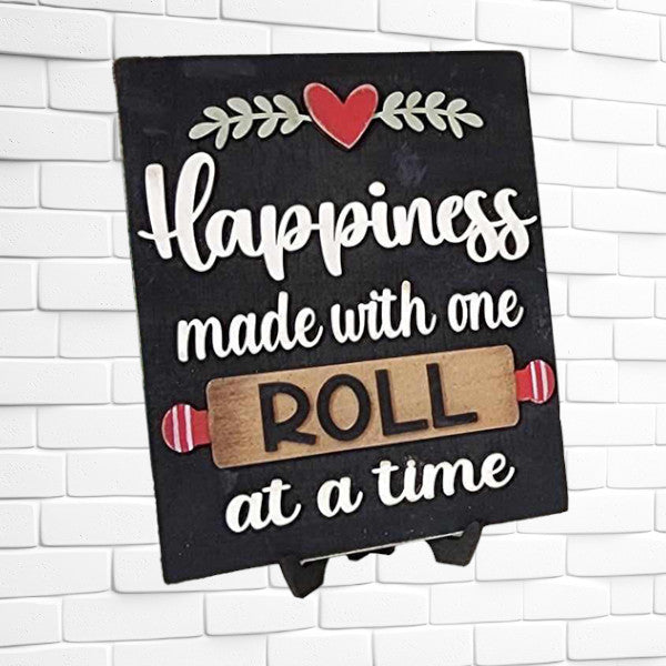 An interchangeable sign from Janet's Craft Corner reads, "Happiness made with one roll at a time," ideal for DIY home decor lovers. The word “ROLL” is accentuated with a rolling pin design, all set against a white brick wall.