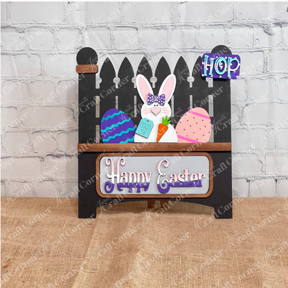 The Happy Easter Bunny Interchangeable Insert by Janet's Craft Corner is a DIY home decor kit featuring an Easter decoration with a black picket fence, a carrot-holding bunny, colorful eggs, and a purple "Happy Easter" sign on burlap against brick. Perfect for holiday-themed decor!.