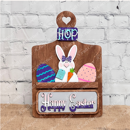 Janet's Craft Corner presents the Easter Bunny Interchangeable Insert, a delightful DIY home decor kit. This wooden decoration showcases a bunny with glasses and a bow tie holding a carrot, accented by colorful eggs and topped with a checkered bow. The "Happy Easter" sign below enhances its charm against a rustic background.
