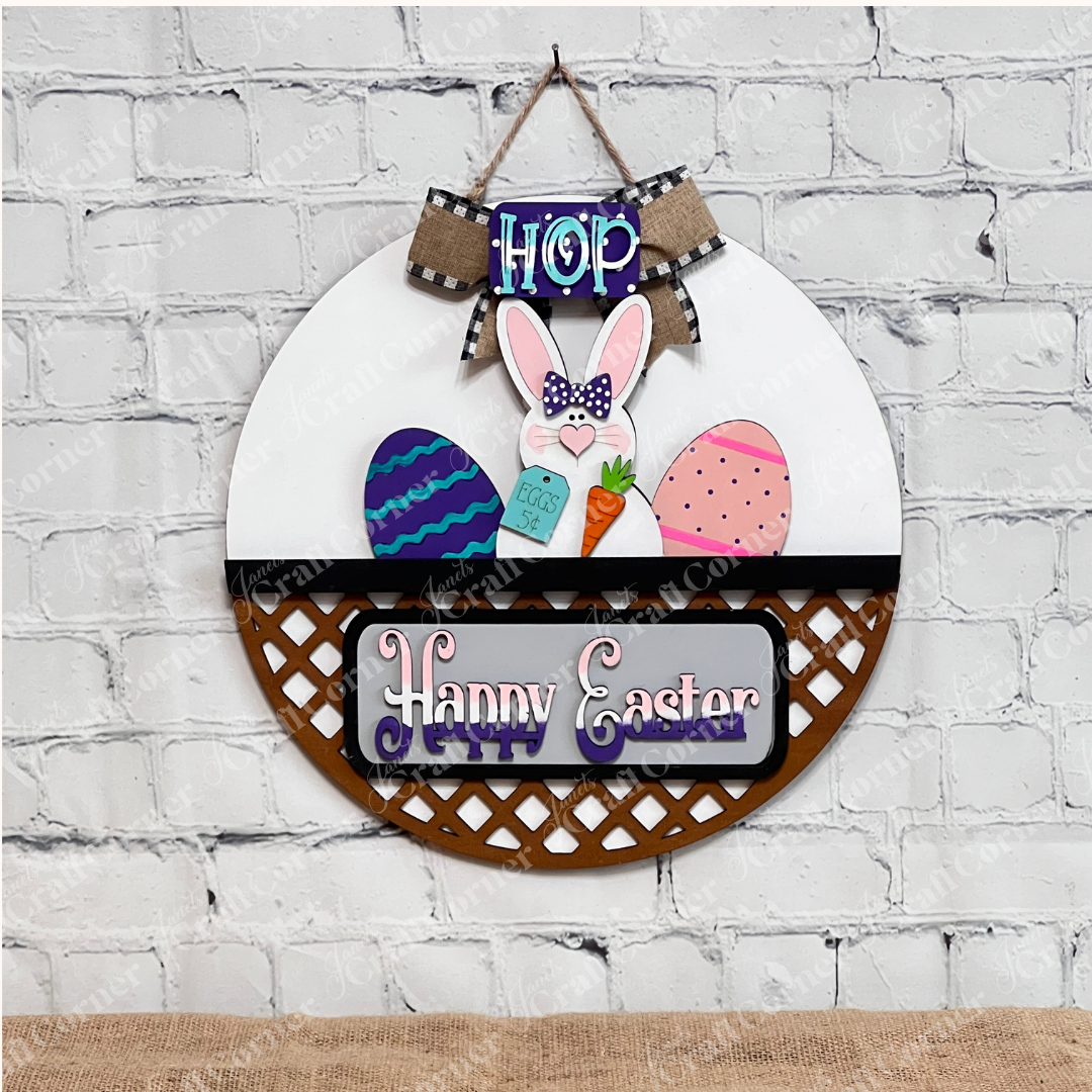 The Janet's Craft Corner Happy Easter Bunny Interchangeable Insert DIY kit features a bunny holding a carrot and colorful eggs, with "Hop" above and "Happy Easter" below. Ideal for home decor, it stands out against a white brick wall background.