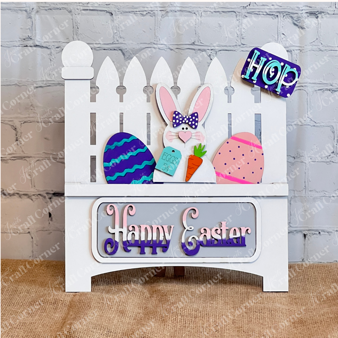 Janet's Craft Corner offers a DIY home decor kit: the Happy Easter Bunny Interchangeable Insert, featuring 3 pieces including a bunny with a carrot and "Eggs 5¢" sign, colorful eggs, and stylized "Happy Easter," perfect for decorating against a brick wall backdrop.