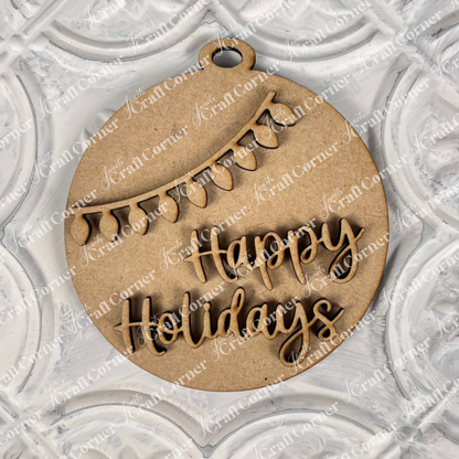 A Round Christmas Ornament from Janet's Craft Corner rests on a textured white surface. This delightful piece, an ideal choice for a DIY decoration kit, showcases cut-out text that says "Happy Holidays" with an elegant string of lights arching above the joyful message.