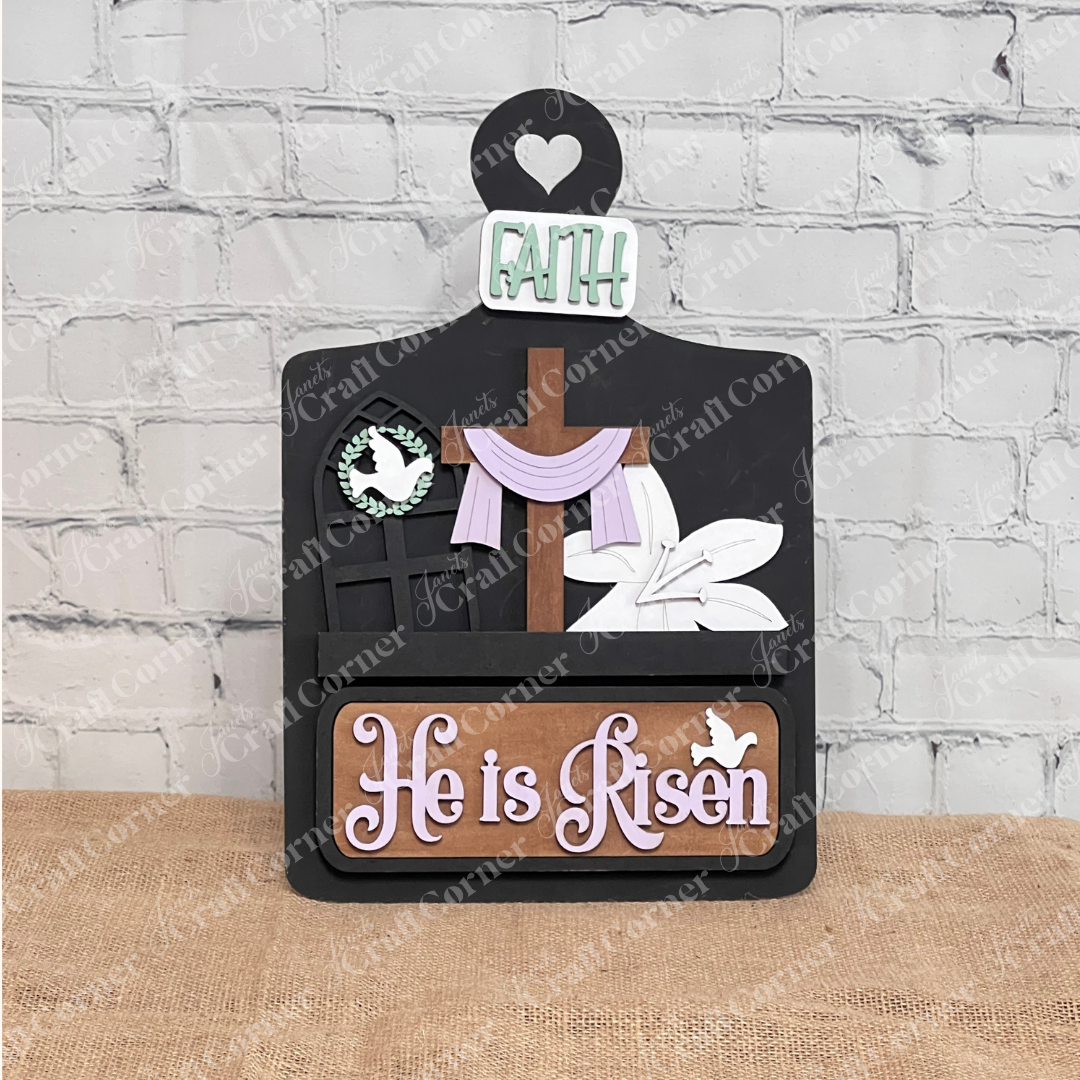 The "He is Risen Interchangeable Insert" DIY craft kit from Janet's Craft Corner includes a wooden sign featuring a cross with fabric, floral touches, a heart cutout, and the words "Faith" and "He is Risen." A dove and window design complete this set of 3 pieces against a white brick wall backdrop.