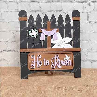 The "He is Risen" Interchangeable Insert from Janet's Craft Corner features a decorative wooden sign with a fence design, adorned with the words in white letters, a cross draped with purple fabric, a white dove, and lilies on burlap against a light brick background for rustic elegance.