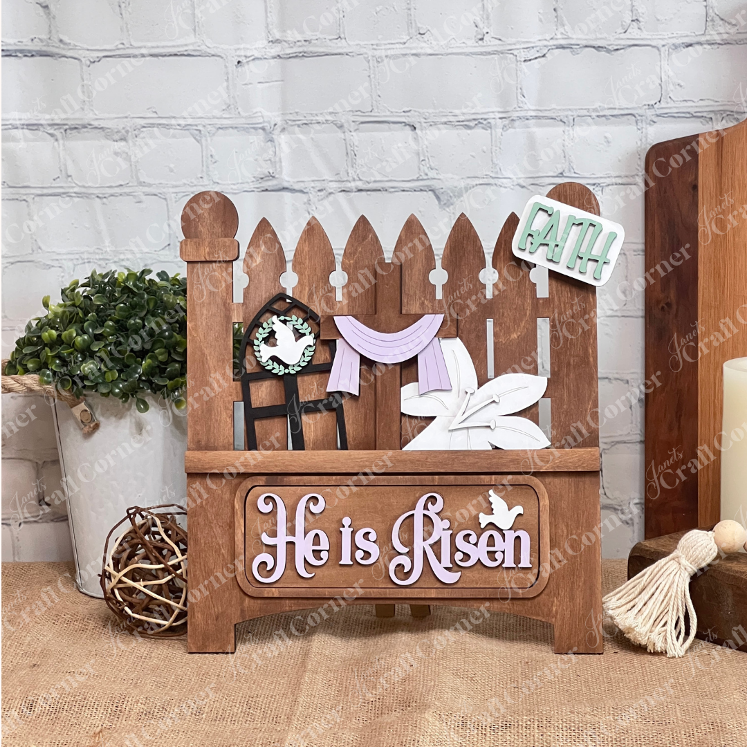 The "He is Risen" interchangeable insert from Janet's Craft Corner is a delightful wooden Easter decoration featuring a fence with a window, a cross draped in purple cloth, a white lily, and "He is Risen" next to a bird silhouette. It includes a green and white "Faith" sign above for versatile home decor.