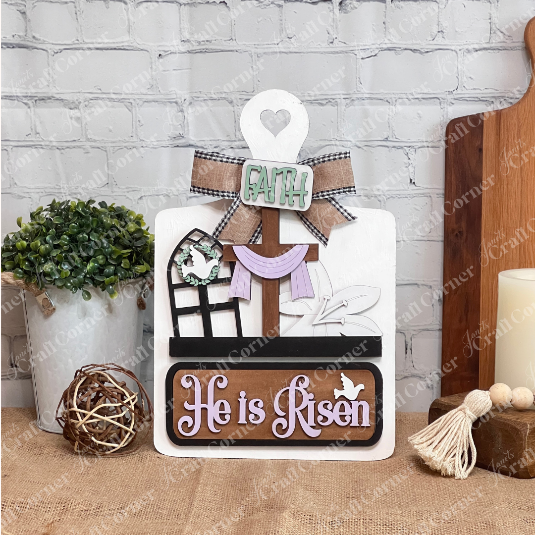 The "He is Risen Interchangeable Insert" DIY craft kit from Janet's Craft Corner includes a wooden sign featuring a cross with fabric, floral touches, a heart cutout, and the words "Faith" and "He is Risen." A dove and window design complete this set of 3 pieces against a white brick wall backdrop.