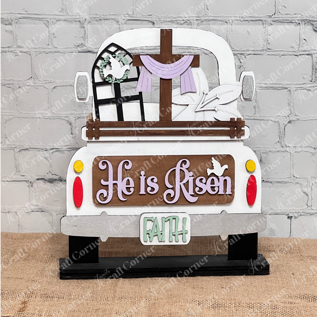 This DIY home decor kit from Janet's Craft Corner includes an interchangeable wooden decoration featuring the back of a truck with "He is Risen" and a dove on the tailgate. It boasts a cross, window frame, cloth in the bed, and "FAITH" on the plate, set against burlap and brick.