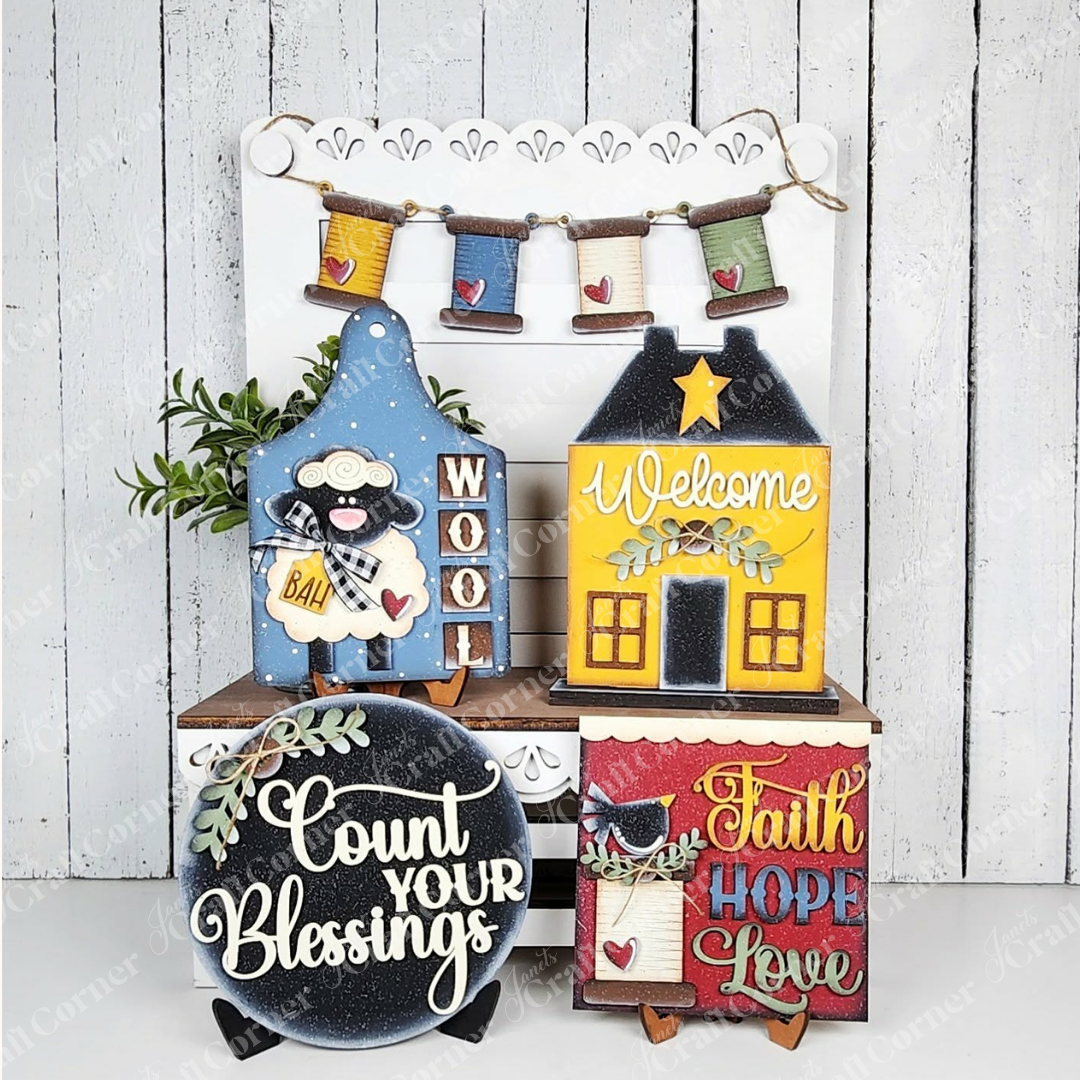 The Heirloom Decor Set from Janet's Craft Corner is a DIY seasonal home decor kit featuring five colorful wood cut-outs: a "Wool" sheep, "Welcome" house, spool garland, "Count Your Blessings" round sign, and cat-themed "Faith Hope Love" square sign.
