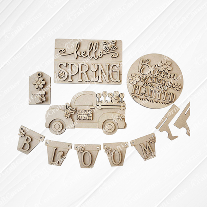 The Hello Spring Decor Set by Janet's Craft Corner is a DIY seasonal home decor craft kit featuring 6 wooden pieces: signs with "hello SPRING" and "Bloom where you are planted," a flower market truck, a small house, and a "BLOOM" banner with delicate flower details.