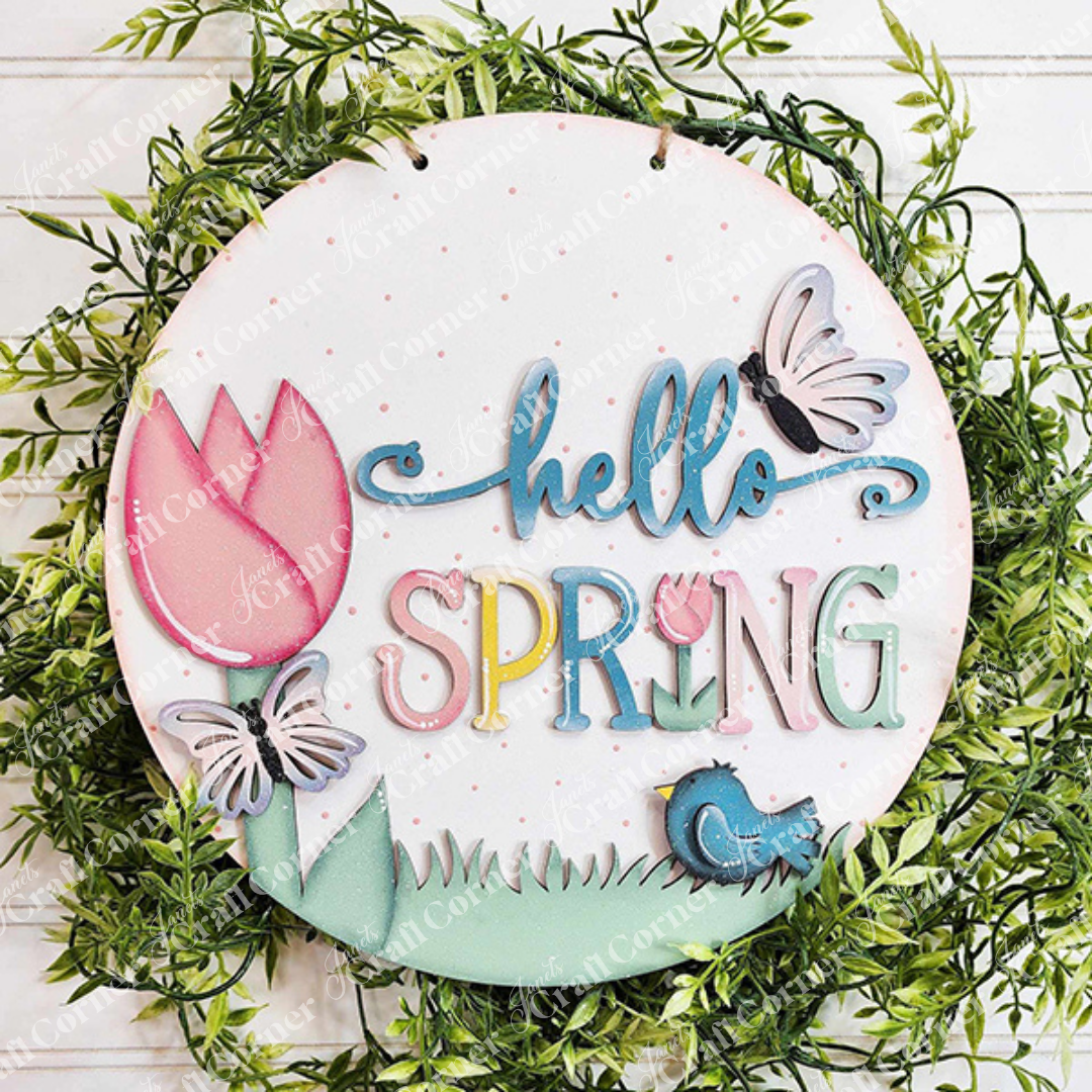 Introducing Janet's Craft Corner's "Hello Spring Round Door Hanger," a charming DIY home decor piece. This delightful door hanger features the cheerful message "hello SPRING" in vibrant letters, accompanied by charming illustrations of a tulip, butterflies, a bird, and lush green leaves. The design is shown attached to a greenery wreath, making it the perfect way to welcome the season.