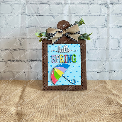 The Spring Switchable Farmhouse Signs DIY kit from Janet's Craft Corner features “hello SPRING,” a colorful umbrella, blue polka dots, faux greenery, and burlap bow—ideal for tiered trays or lavender decor on a burlap backdrop.