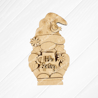 The Hellp Spring Girl Gnome Shelf Sitter by Janet's Craft Corner, a DIY seasonal home decor craft kit, features a wooden girl gnome with a floral hat holding a "Hello Spring" mug. It's adorned with flowers and a small bird against a textured white background.