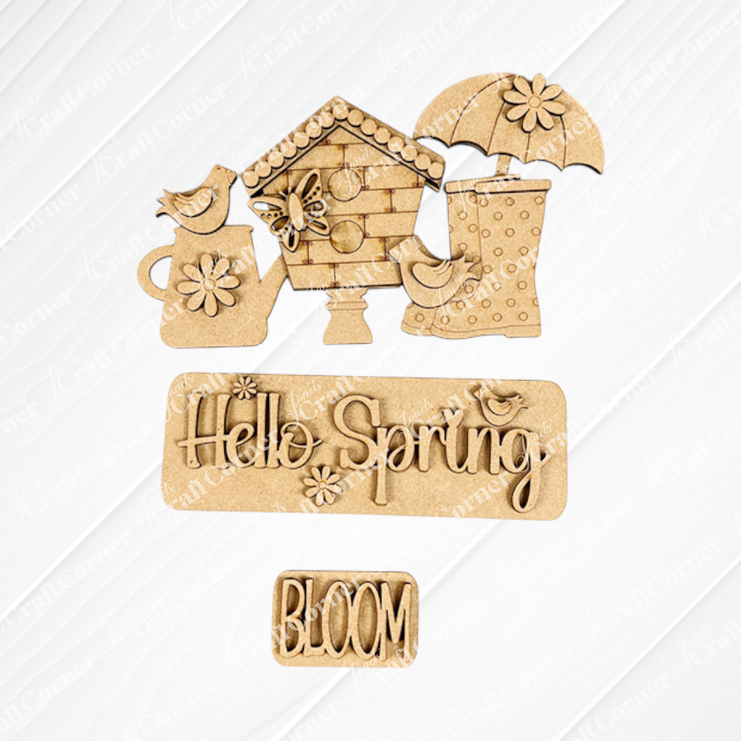 Janet's Craft Corner presents the "Hello Spring Interchangeable Insert" DIY home decor kit, featuring wooden cutouts of a birdhouse, birds, an umbrella, boots, and flowers on a textured background. The text reads "Hello Spring" and "BLOOM," making it perfect for crafting your own springtime decorative insert!