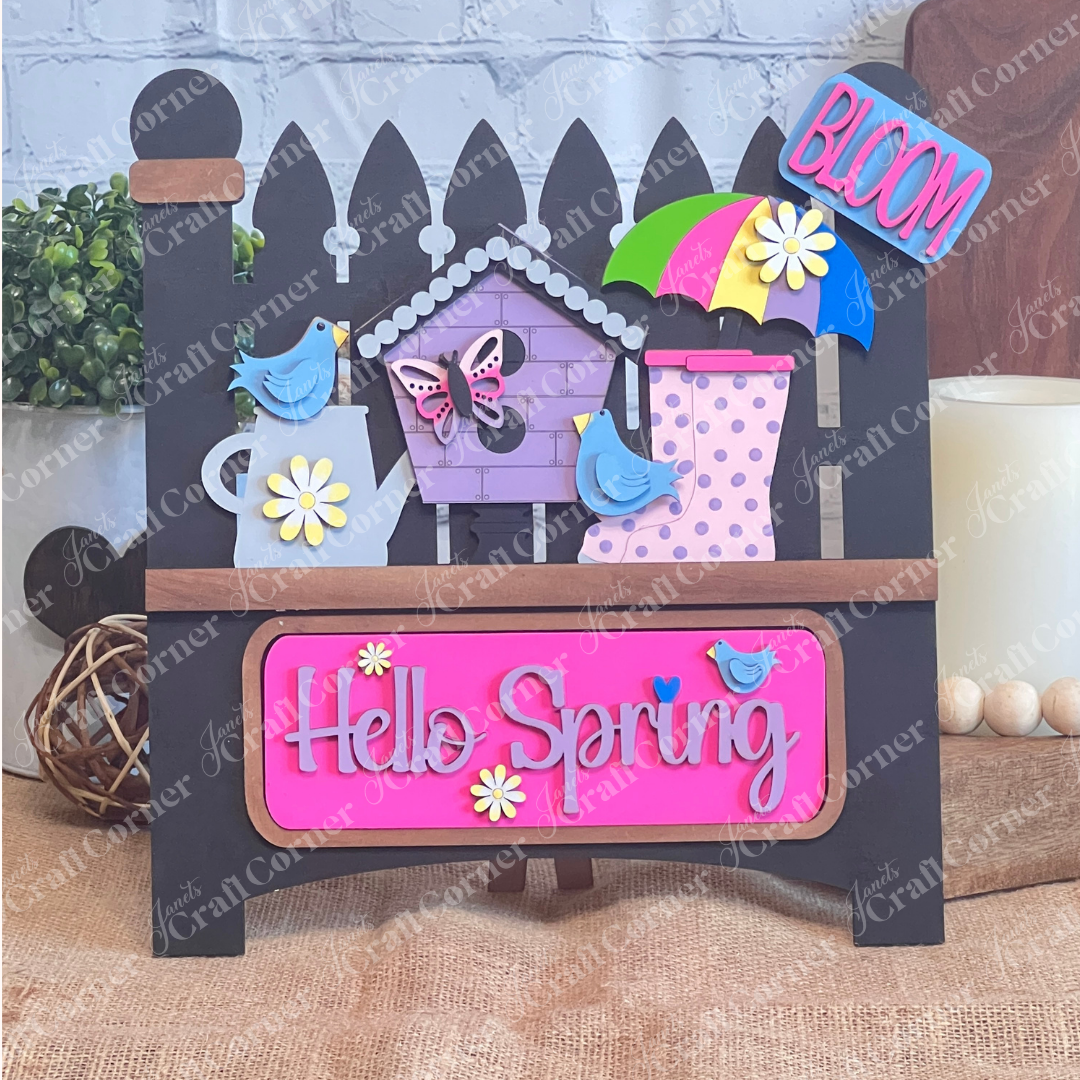 A vibrant wooden decoration from Janet's Craft Corner featuring the "Hello Spring" interchangeable insert. This DIY home decor kit showcases a black garden fence adorned with the phrases "Hello Spring" and "Bloom." It's embellished with flowers, a butterfly, birds, an umbrella, and a birdhouse, perfectly capturing the essence of spring.