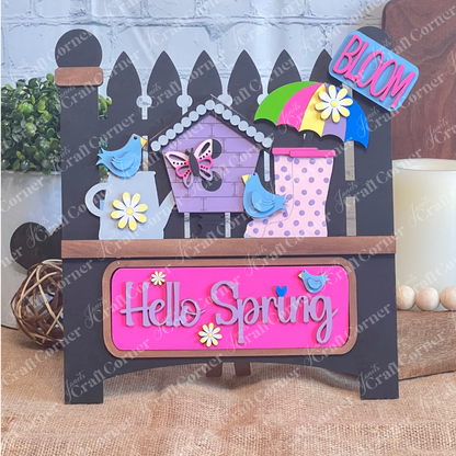 A vibrant wooden decoration from Janet's Craft Corner featuring the "Hello Spring" interchangeable insert. This DIY home decor kit showcases a black garden fence adorned with the phrases "Hello Spring" and "Bloom." It's embellished with flowers, a butterfly, birds, an umbrella, and a birdhouse, perfectly capturing the essence of spring.