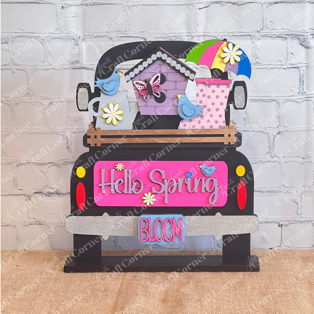 A vibrant wooden decoration from Janet's Craft Corner featuring the "Hello Spring" interchangeable insert. This DIY home decor kit showcases a black antique truck decor adorned with the phrases "Hello Spring" and "Bloom." It's embellished with flowers, a butterfly, birds, an umbrella, and a birdhouse, perfectly capturing the essence of spring.