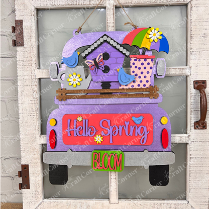 A vibrant wooden decoration from Janet's Craft Corner hangs on a rustic white door, featuring the "Hello Spring" interchangeable insert. This DIY home decor kit showcases a purple truck adorned with the phrases "Hello Spring" and "Bloom." It's embellished with flowers, a butterfly, birds, an umbrella, and a birdhouse, perfectly capturing the essence of spring.
