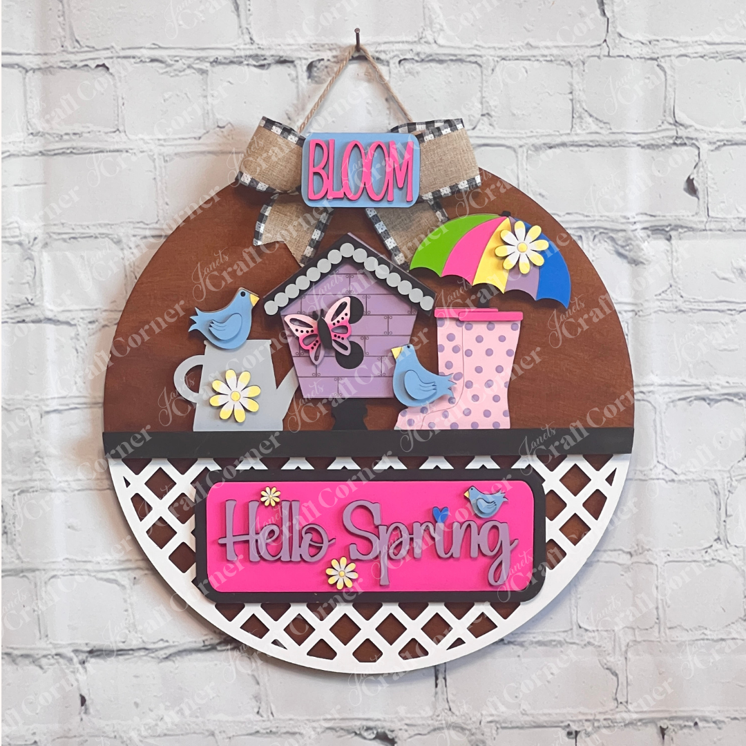 A vibrant wooden decoration from Janet's Craft Corner featuring the "Hello Spring" interchangeable insert. This DIY home decor kit showcases a stained door hanger adorned with the phrases "Hello Spring" and "Bloom." It's embellished with flowers, a butterfly, birds, an umbrella, and a birdhouse, perfectly capturing the essence of spring.