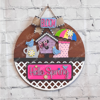 A vibrant wooden decoration from Janet's Craft Corner featuring the "Hello Spring" interchangeable insert. This DIY home decor kit showcases a stained door hanger adorned with the phrases "Hello Spring" and "Bloom." It's embellished with flowers, a butterfly, birds, an umbrella, and a birdhouse, perfectly capturing the essence of spring.