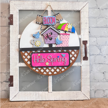 A vibrant wooden decoration from Janet's Craft Corner featuring the "Hello Spring" interchangeable insert. This DIY home decor kit showcases a white door hanger adorned with the phrases "Hello Spring" and "Bloom." It's embellished with flowers, a butterfly, birds, an umbrella, and a birdhouse, perfectly capturing the essence of spring.