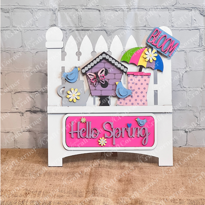 A vibrant wooden decoration from Janet's Craft Corner featuring the "Hello Spring" interchangeable insert. This DIY home decor kit showcases a white garden fence adorned with the phrases "Hello Spring" and "Bloom." It's embellished with flowers, a butterfly, birds, an umbrella, and a birdhouse, perfectly capturing the essence of spring.