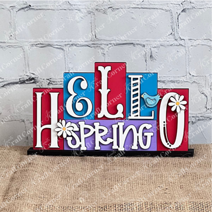The Hello Spring Mini Word Block from Janet's Craft Corner features vibrant wooden lettering, with playful font variations. Ideal for home décor, it showcases a charming blue bird perched on the letter "R," and delightful daisies embellishing the "H" and "O." Positioned against a white brick wall on a burlap surface, it's perfect for adding a touch of spring to any room.