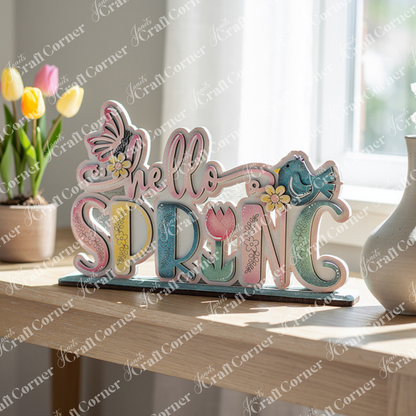 Introducing the Hello Spring Standing Shelf Sitter by Janet's Craft Corner. This delightful DIY kit features a decorative wooden sign with "hello SPRING" in pastel colors, complete with charming butterfly and bird accents perched on the letters and adorned with flower designs. Perfect for home décor enthusiasts, it makes an eye-catching display on a wooden table next to a tulip potted plant.