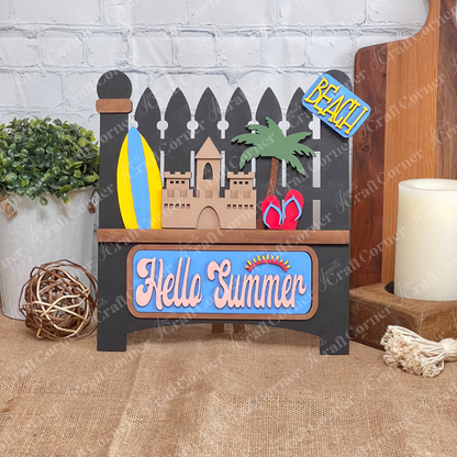 Introducing the Summer Beach Interchangeable Insert, a DIY home decor kit from Janet's Craft Corner. This wooden summer-themed decoration showcases a delightful sandcastle, surfboards, and palm trees, all sitting on a black garden fence. The sign displays "Hello Summer" in colorful letters, making it an ideal piece for adding a vibrant touch to your space.