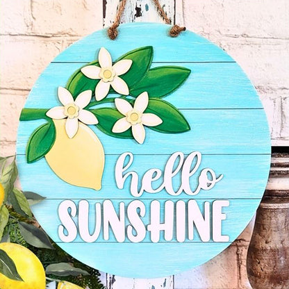 The Hello Sunshine Lemon Round Sign by Janet's Craft Corner showcases a painted lemon and white flowers on a delightful teal background. The phrase "hello SUNSHINE" in white letters brings a cheerful element to your home décor, making it an ideal piece to hang against a brick wall and brighten any space.