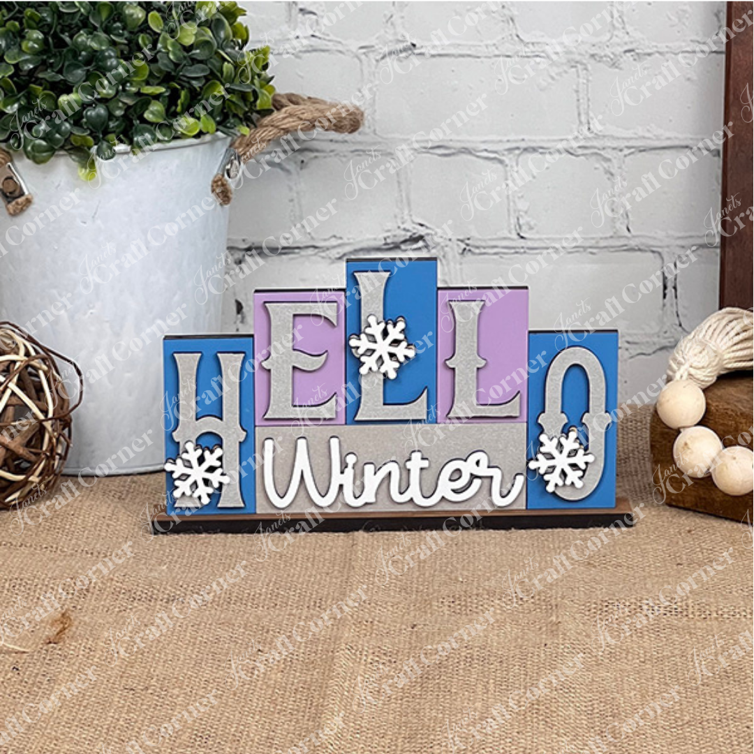 Introducing the "Hello Winter" Mini Word Block Shelf Sitter by Janet's Craft Corner. This DIY decor kit features blue, purple, and gray blocks with snowflake-adorned letters. It is showcased on a burlap-covered surface, complete with greenery and wooden beads, making it an ideal addition to your home décor collection.