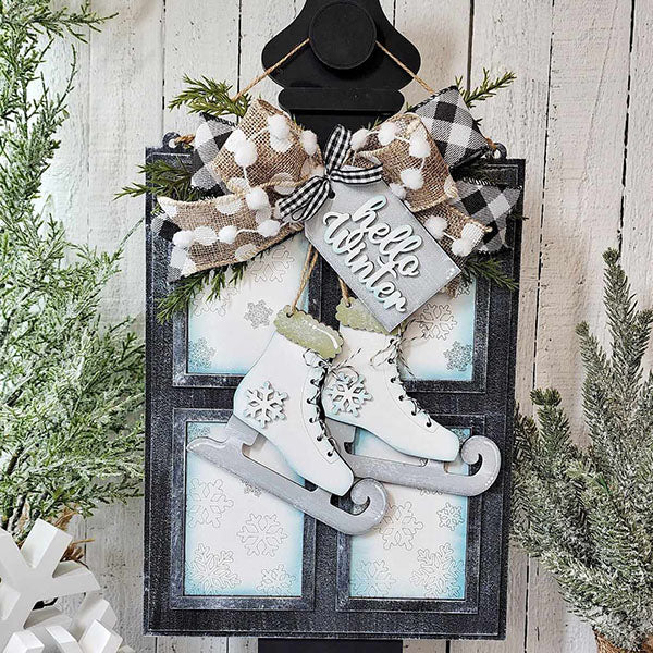 The "Hello Winter Window Hanger" from Janet's Craft Corner showcases festive winter decor, including ice skates and a welcoming "hello winter" sign, enhanced by pine branches and a black and white gingham bow. A snowflake-decorated white wooden background adds the finishing touch, making it ideal for home décor this season.