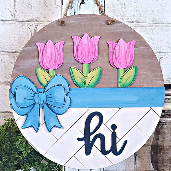 The "Hi Tulip Round" from Janet's Craft Corner is a delightful DIY kit that showcases a round wooden sign embellished with three pink tulips, a large blue bow, and the word "hi" at the bottom. Its elegant gray and white diagonal background and rope for hanging make it a charming addition to any home décor collection.