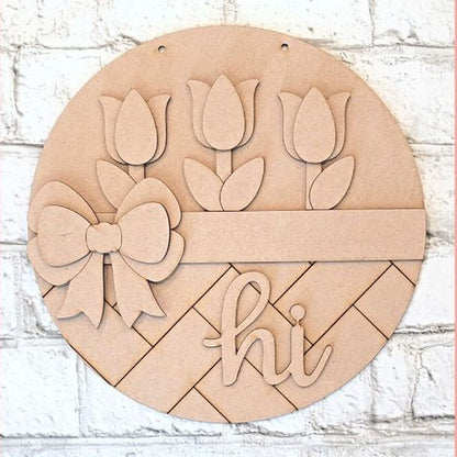 The Hi Tulip Round from Janet's Craft Corner is a DIY seasonal home decor craft kit featuring a round wooden plaque with tulip cut-outs, a large bow, and "hi" at the bottom, perfect for enhancing any space against a white brick wall.