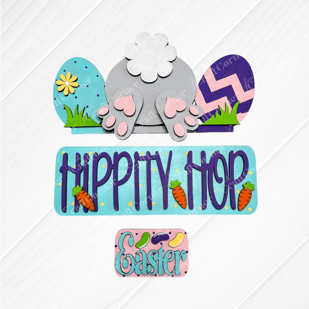The Hippity Hop Bunny Butt Interchangeable Insert from Janet's Craft Corner is a charming DIY craft kit featuring a bunny tail, colorful eggs, paw prints, and "Hippity Hop" in blue with carrot accents on  a white wooden background.