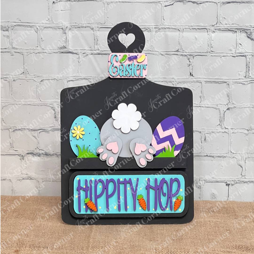 The "Hippity Hop Bunny Butt Interchangeable Insert" by Janet's Craft Corner features a decorative bunny rear with Easter eggs. The "Hippity Hop" sign is perfect home decor against a white brick wall for a festive touch.