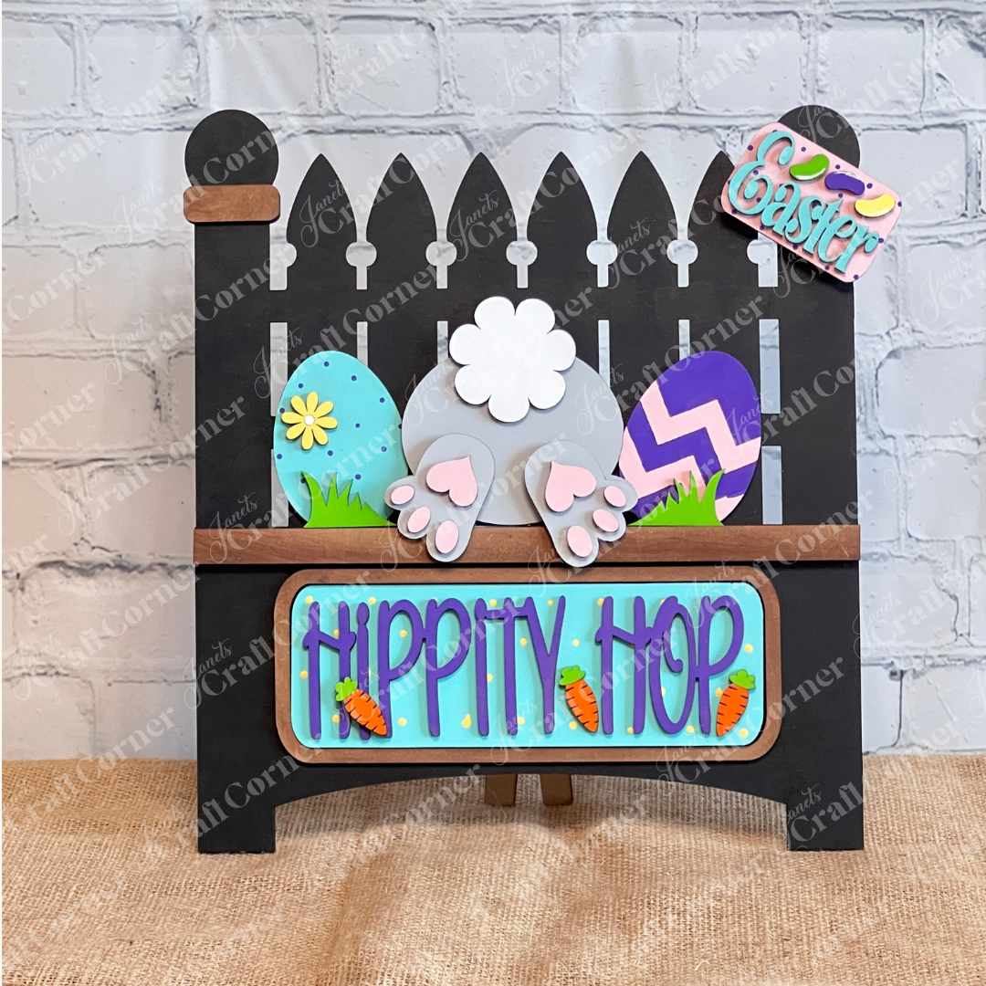 The Hippity Hop Bunny Butt Interchangeable Insert from Janet's Craft Corner is a charming DIY craft kit featuring a decorative black picket fence sign with a bunny tail, colorful eggs, paw prints, and "Hippity Hop" in blue with carrot accents on burlap against a white brick wall.