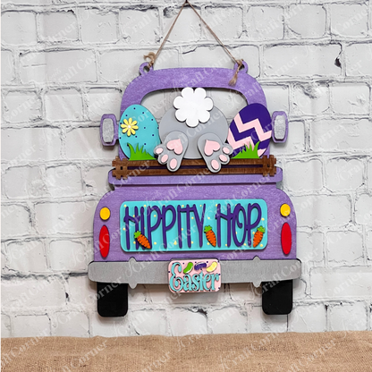 The "Hippity Hop Bunny Butt Interchangeable Insert" from Janet's Craft Corner features a hand-painted purple truck filled with Easter eggs and a bunny's hindquarters, set against a white brick background. It includes the text "Hippity Hop" with an "Easter" sign below, ideal for home decor.