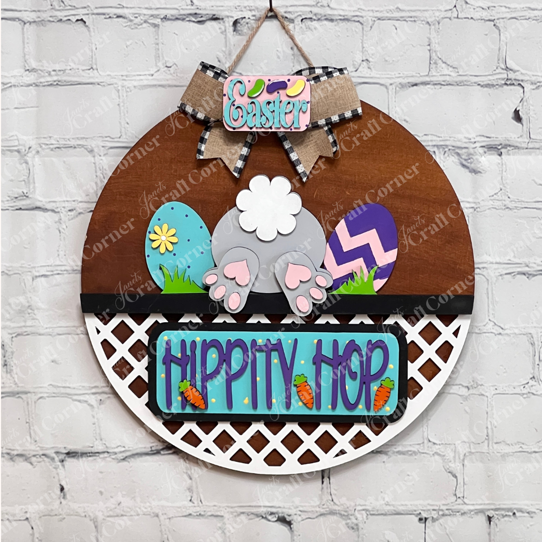 The DIY craft kit "Hippity Hop Bunny Butt Interchangeable Insert" by Janet's Craft Corner features a hand-painted wooden bunny's back, surrounded by colorful eggs and small carrots. The sign reads "Hippity Hop," and it hangs on a brick wall with an "Easter" bow at the top.