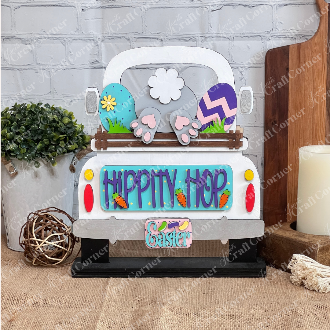 The "Hippity Hop Bunny Butt Interchangeable Insert" by Janet's Craft Corner is a decorative sign featuring a hand-painted bunny tail and feet in a truck bed, surrounded by Easter eggs, flowers, and carrot decorations against a brick wall backdrop for versatile home decor.