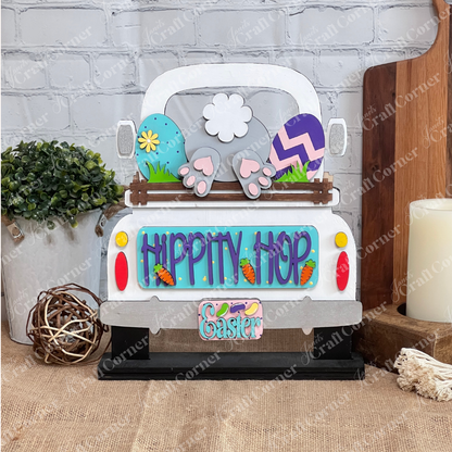 The "Hippity Hop Bunny Butt Interchangeable Insert" by Janet's Craft Corner is a decorative sign featuring a hand-painted bunny tail and feet in a truck bed, surrounded by Easter eggs, flowers, and carrot decorations against a brick wall backdrop for versatile home decor.