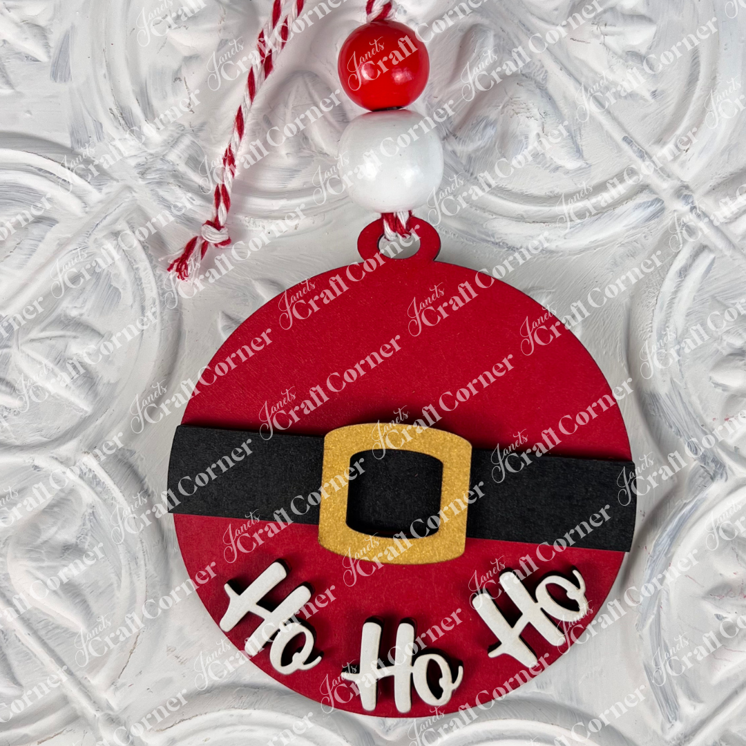 Introducing Janet's Craft Corner's Round Christmas Ornaments from the DIY Decoration Kits collection. This ornament features a vibrant red design with a black belt and gold buckle, reminiscent of Santa's iconic outfit. The phrase "Ho Ho Ho" is elegantly scripted in white below the belt, making it an ideal choice for infusing festive spirit into your home décor. It hangs gracefully from a string decorated with red and white beads, set against a textured background.