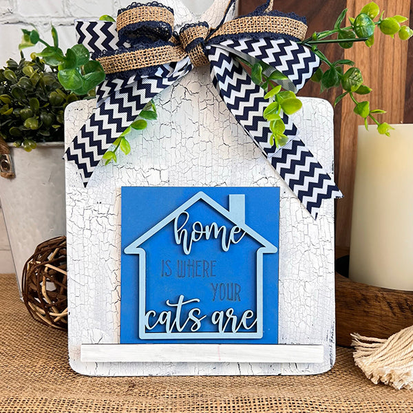 The Cat Swappable Squares by Janet's Craft Corner is a blue house-shaped decorative sign with "Home is where your cats are," a black-and-white chevron ribbon, and greenery on a rustic white background, perfect for tiered trays.