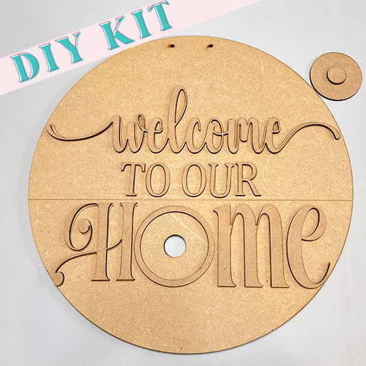 Introducing the Welcome to Our Home Door Hanger Interchangeable Set by Janet's Craft Corner. This round wooden DIY kit features cut-out letters that spell "Welcome to Our Home" against a light backdrop, with a smaller circular piece included for added versatility in your home décor. The top left proudly displays the label "DIY KIT.